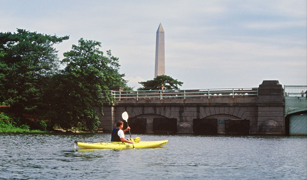 A man in a yellow kayak paddles down the river with the washington monument visible in the distance.