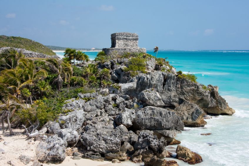 One of the iconic structures of Tulum rises above rocky coastline with a beach visible in the foreground.